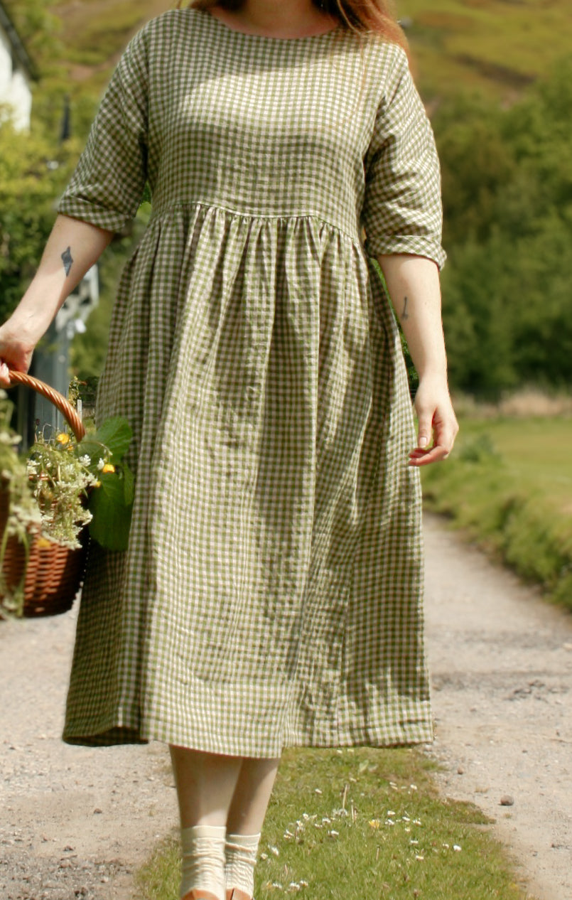 Woman in a green checkered dress standing on a path with a basket, surrounded by nature.