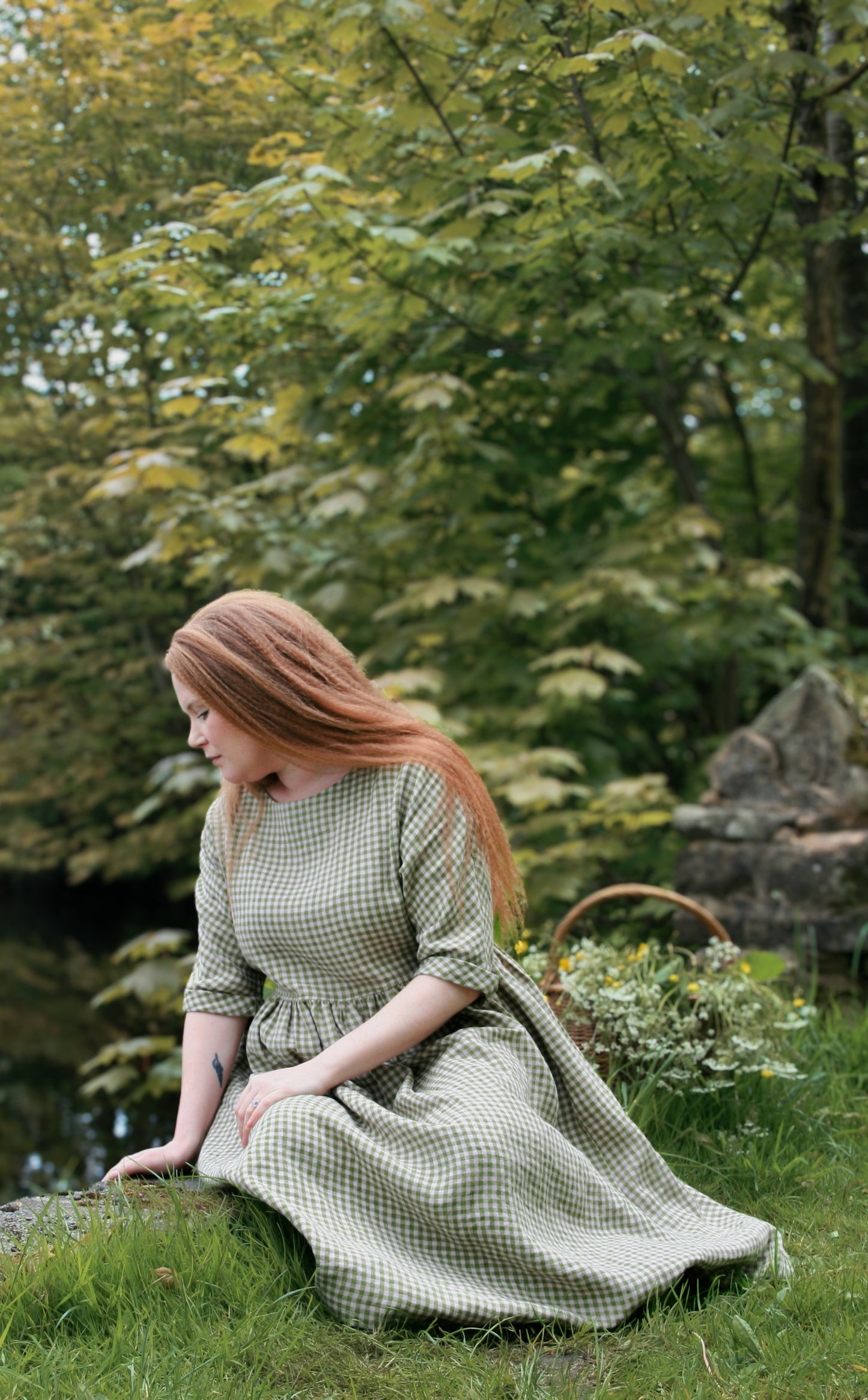 Woman in a green checkered dress sitting on grass with trees in the background