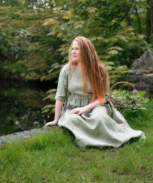 Woman in a green checkered dress sitting on grass with trees in the background