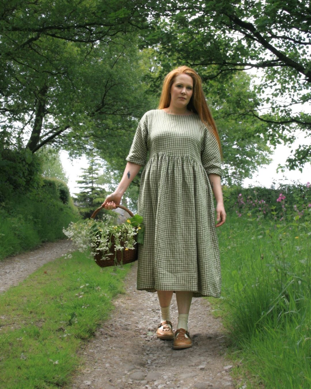 Woman in a green checkered dress holding a basket of plants in a garden path.