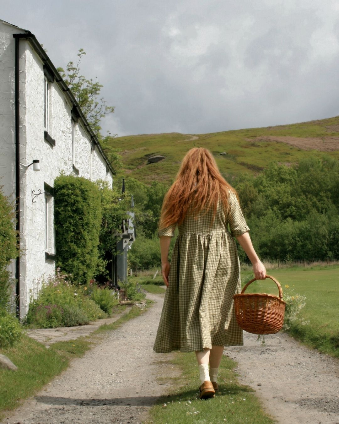 Woman with long red hair in a green dress walking away from a white building towards a grassy hill.