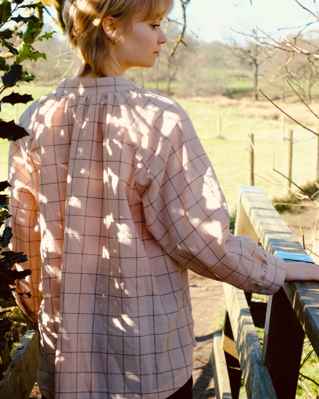 Person wearing a pink plaid shirt standing outdoors with sunlight filtering through leaves.