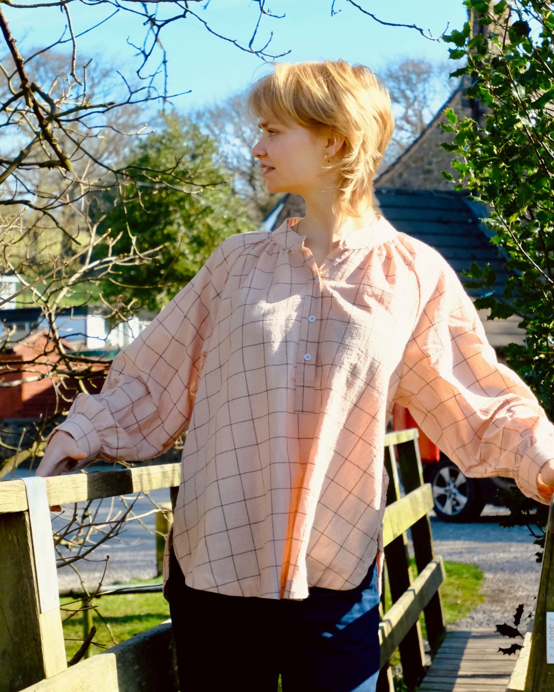 Person wearing a check shirt standing outdoors with trees and a house in the background