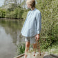 Woman standing by a lake holding a straw bag, wearing a light blue shirt and white shorts.