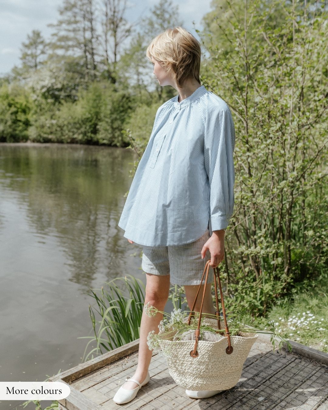 Woman standing by a lake holding a straw bag, wearing a light blue shirt and white shorts.