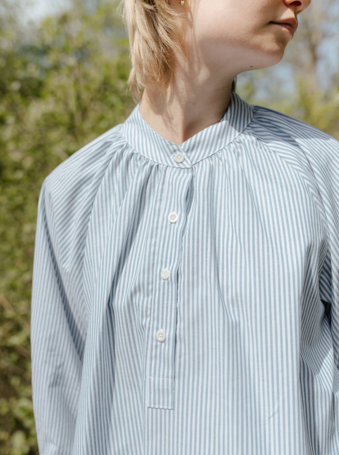 Person wearing a light blue and white striped shirt outdoors with greenery in the background