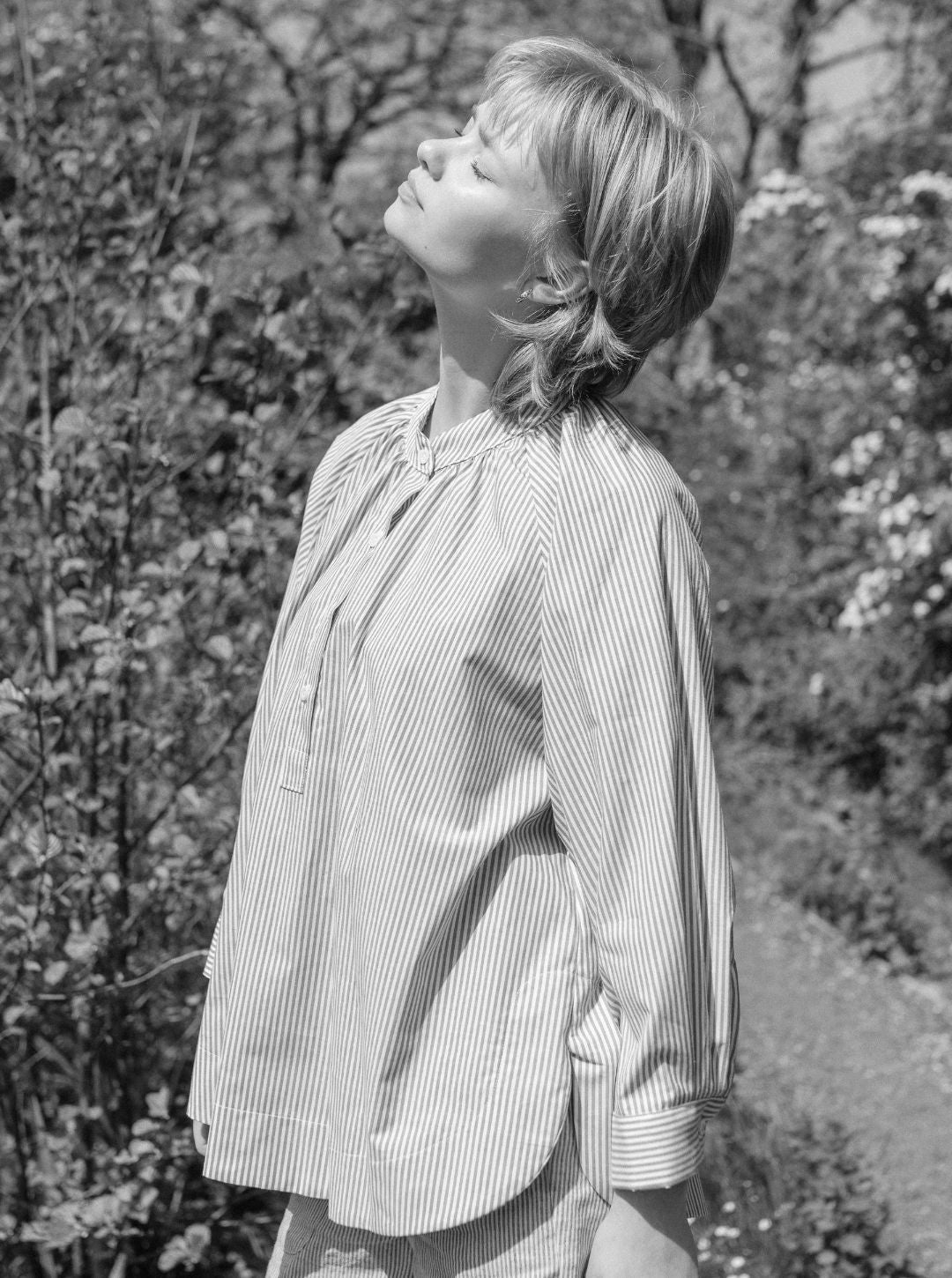 Person wearing a striped shirt standing outdoors with trees and flowers in the background