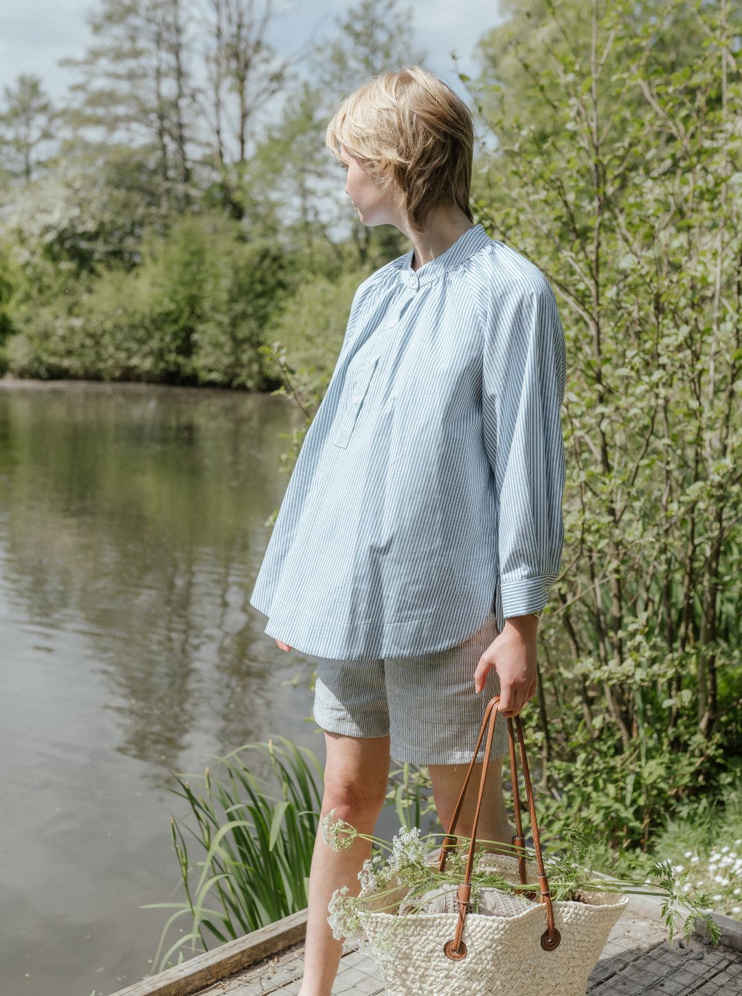 Woman standing by a lake wearing a light blue shirt and gray shorts, holding a straw bag.