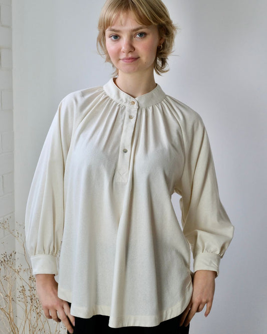 A woman wearing a beige, long-sleeved, smock-style blouse with a half placket and round neckline.