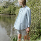 Woman standing by a lake wearing a light blue shirt and gray shorts, holding a straw bag.