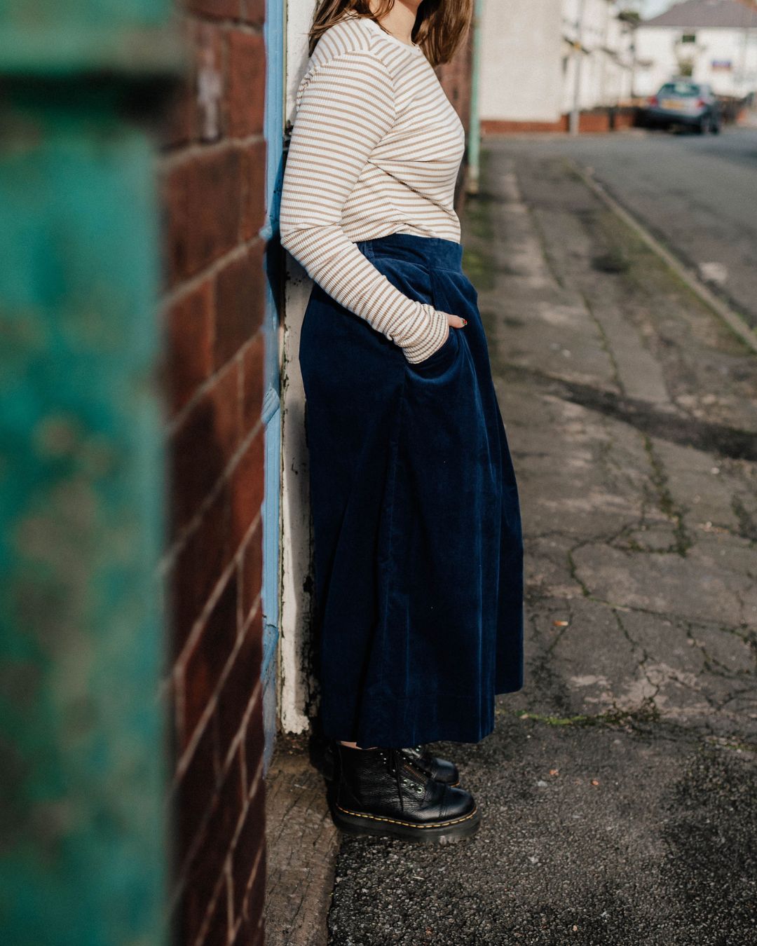 Person wearing a striped sweater and navy skirt standing against a wall on a street.