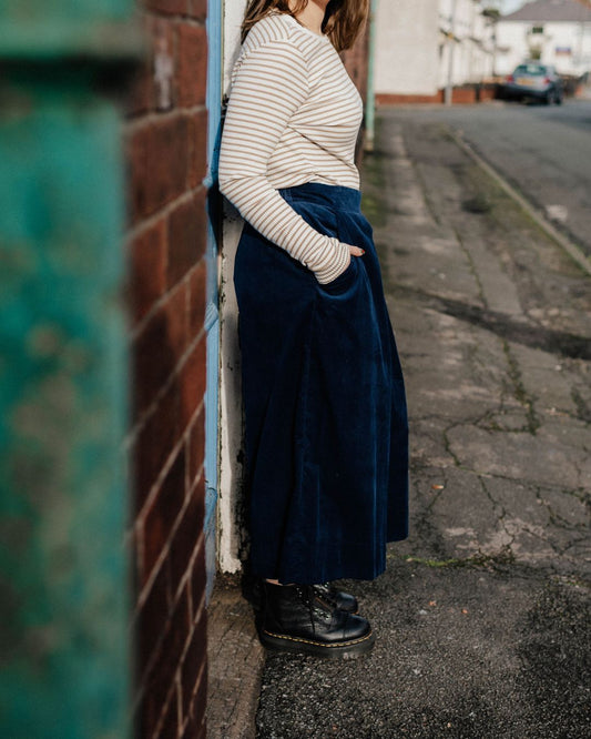 Person wearing a striped sweater and navy skirt standing against a wall on a street.