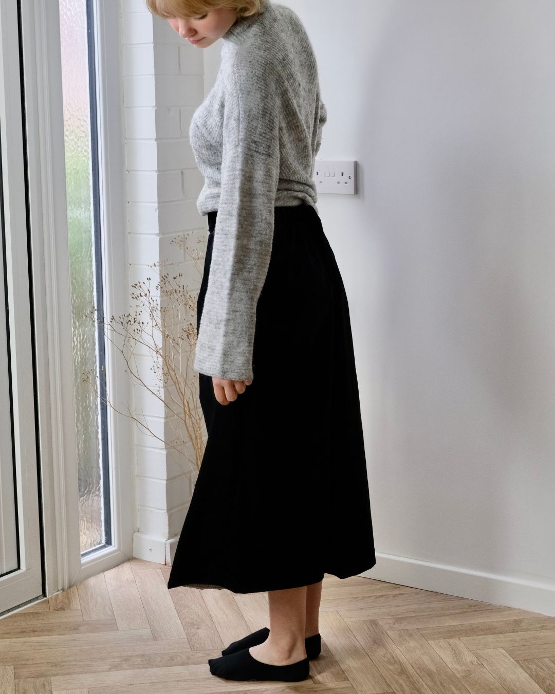 woman standing in studio looking down wearing grey jumper and black clotted
