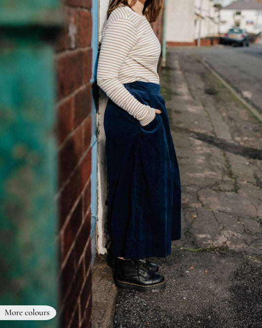 Person wearing a striped sweater and navy skirt standing against a wall on a street.