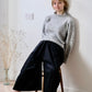 Person wearing a gray sweater and black culottes sitting on a wooden stool against a white wall.