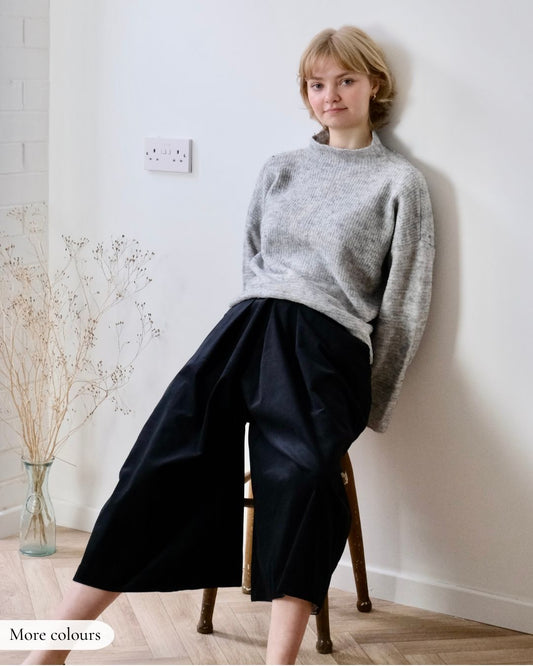 Person wearing a gray sweater and black culottes sitting on a wooden stool against a white wall.