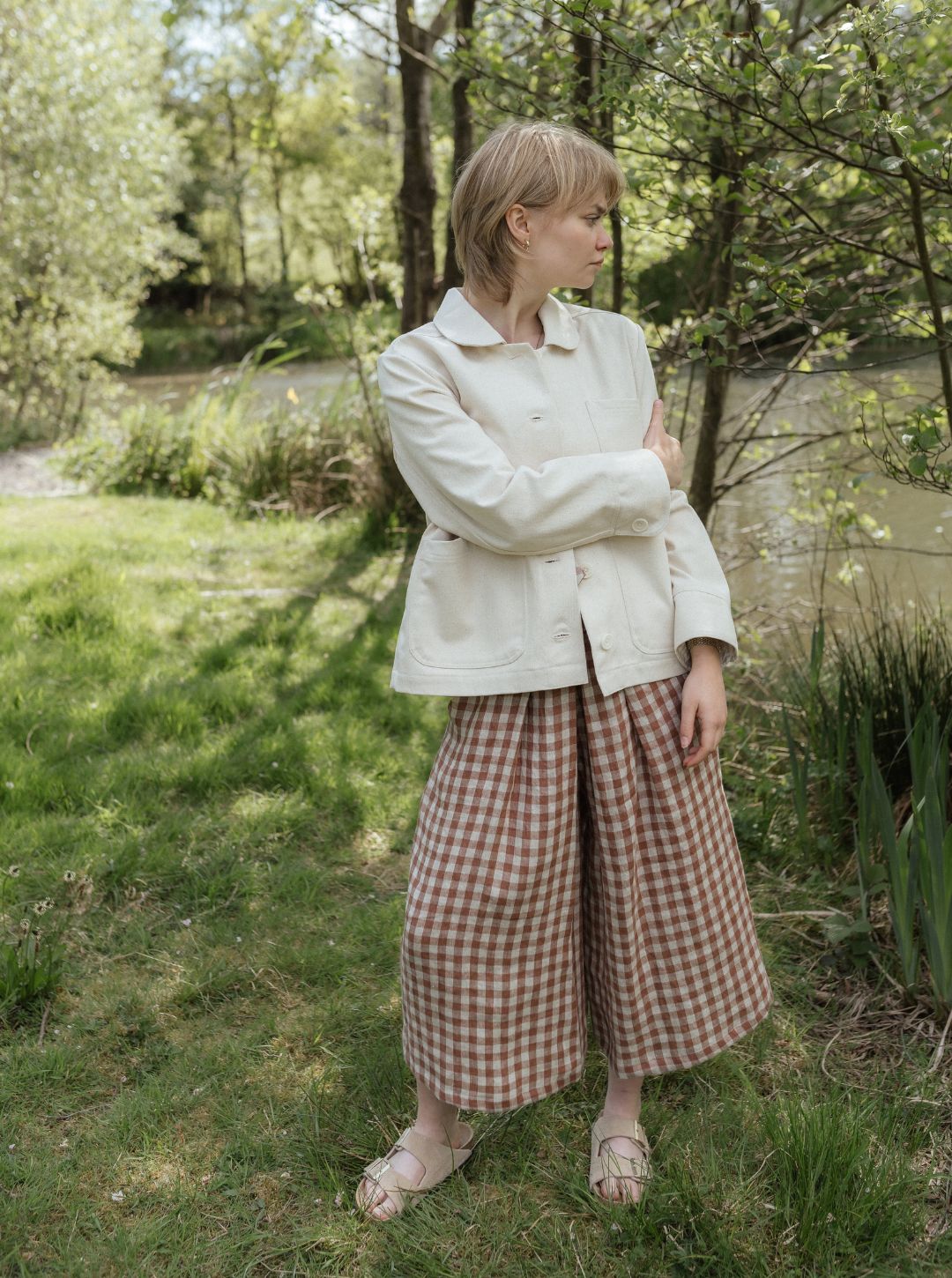 Woman in a white jacket and checkered pants standing in a natural setting with trees and grass.