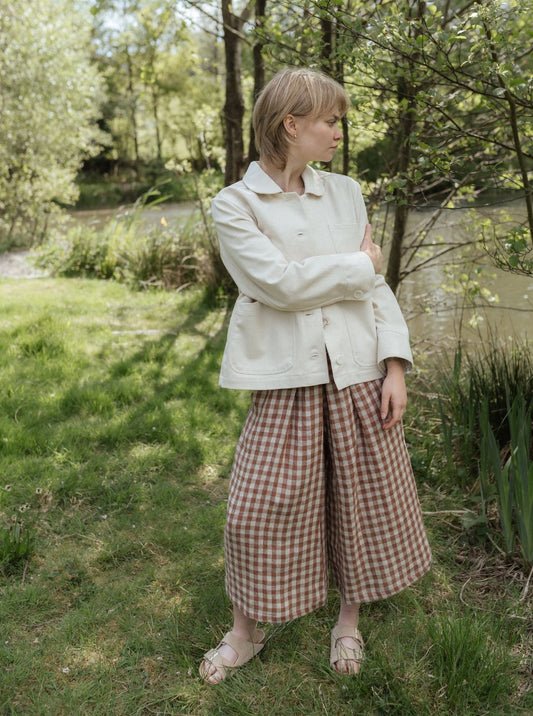 Woman in a white jacket and checkered pants standing in a natural setting with trees and grass.