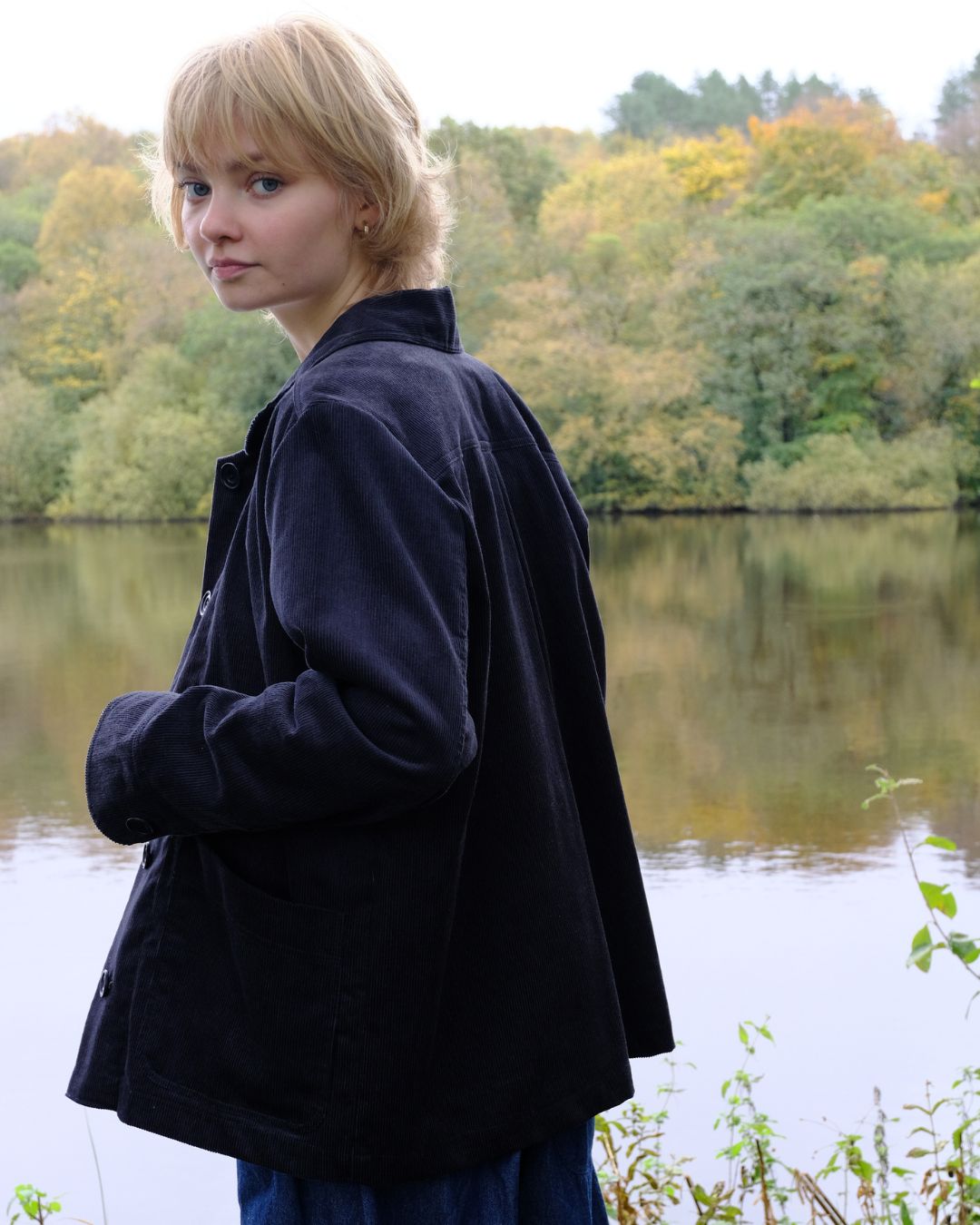 Woman in a dark coat standing by a lake with trees in the background