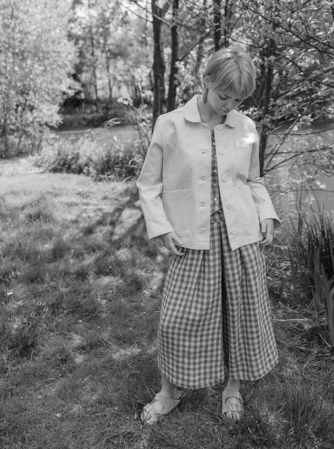 Person wearing a light jacket and checkered skirt standing in a garden with trees and grass.