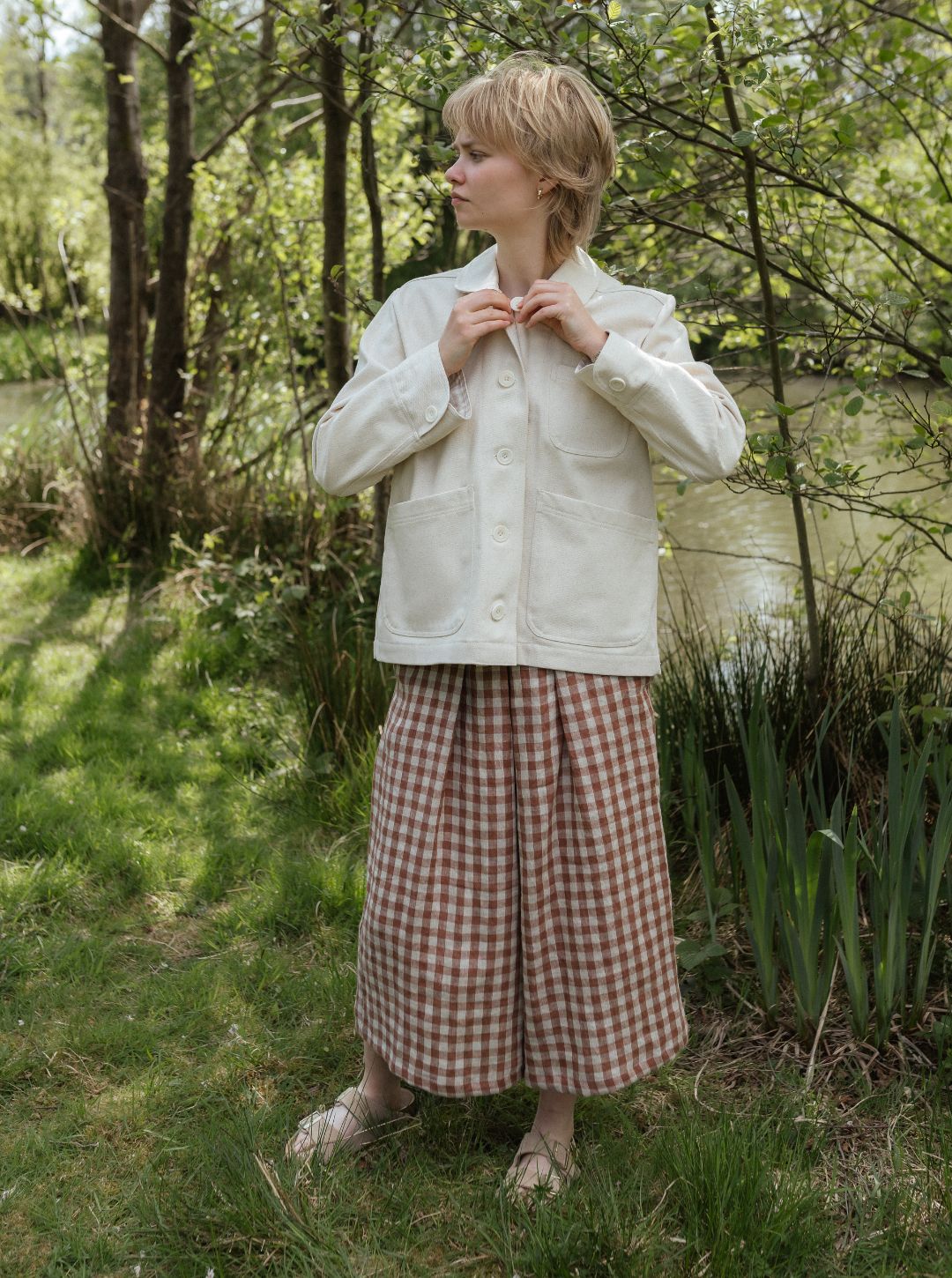 Woman in a white jacket and checkered skirt standing in a natural setting with trees and water.