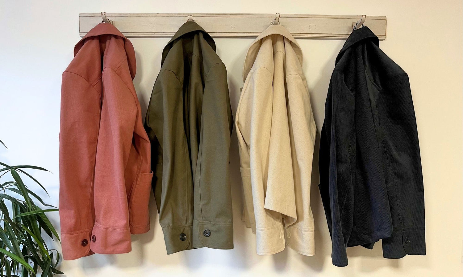 Four jackets hanging on a rack against a white wall with a plant to the left.