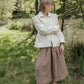 Woman in a white jacket and checkered pants standing in a natural setting with trees and grass.