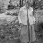 Person wearing a light jacket and checkered skirt standing in a garden with trees and grass.
