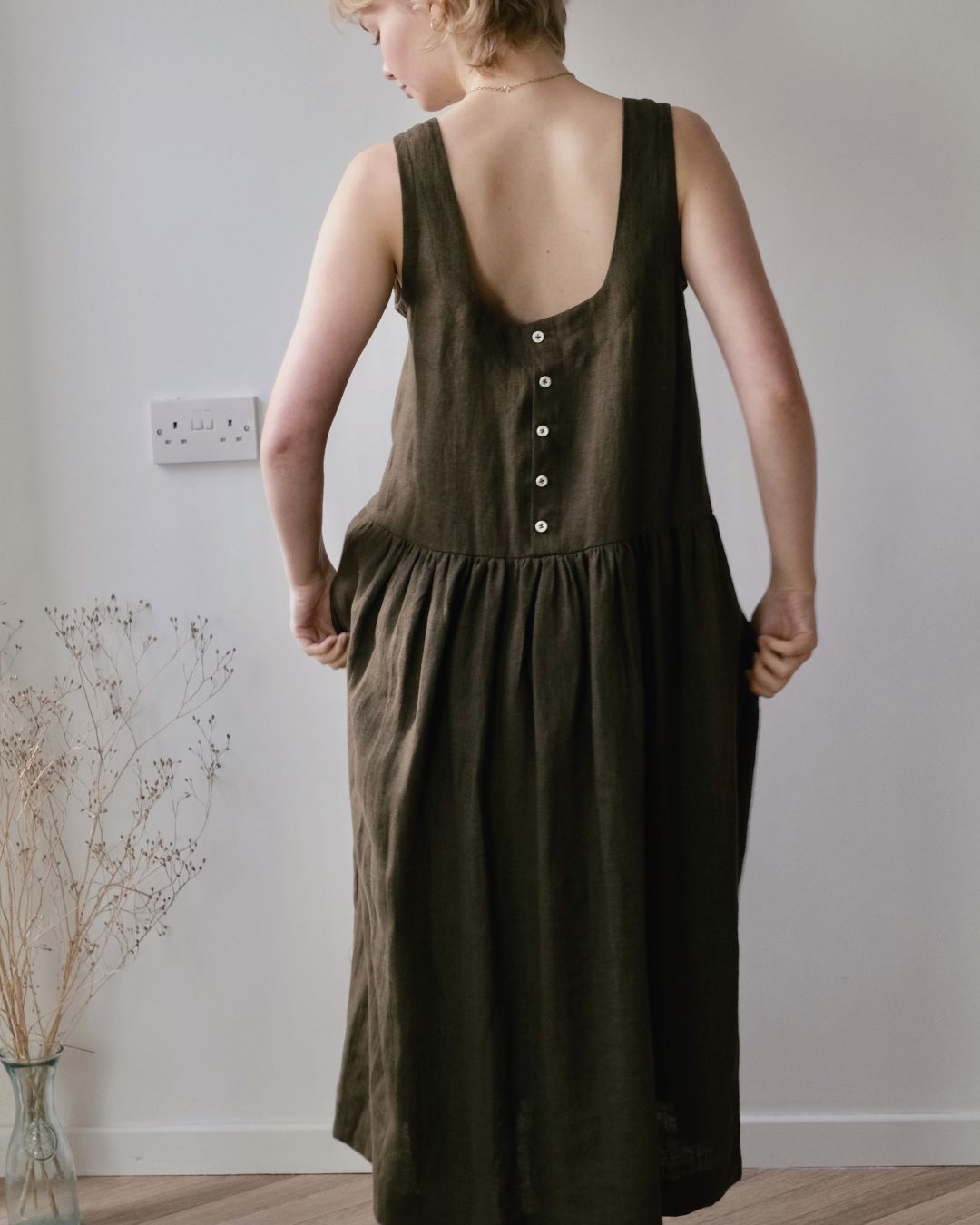 back view of woman standing in conservatory wearing sleeveless pinafore dress with buttoned back.