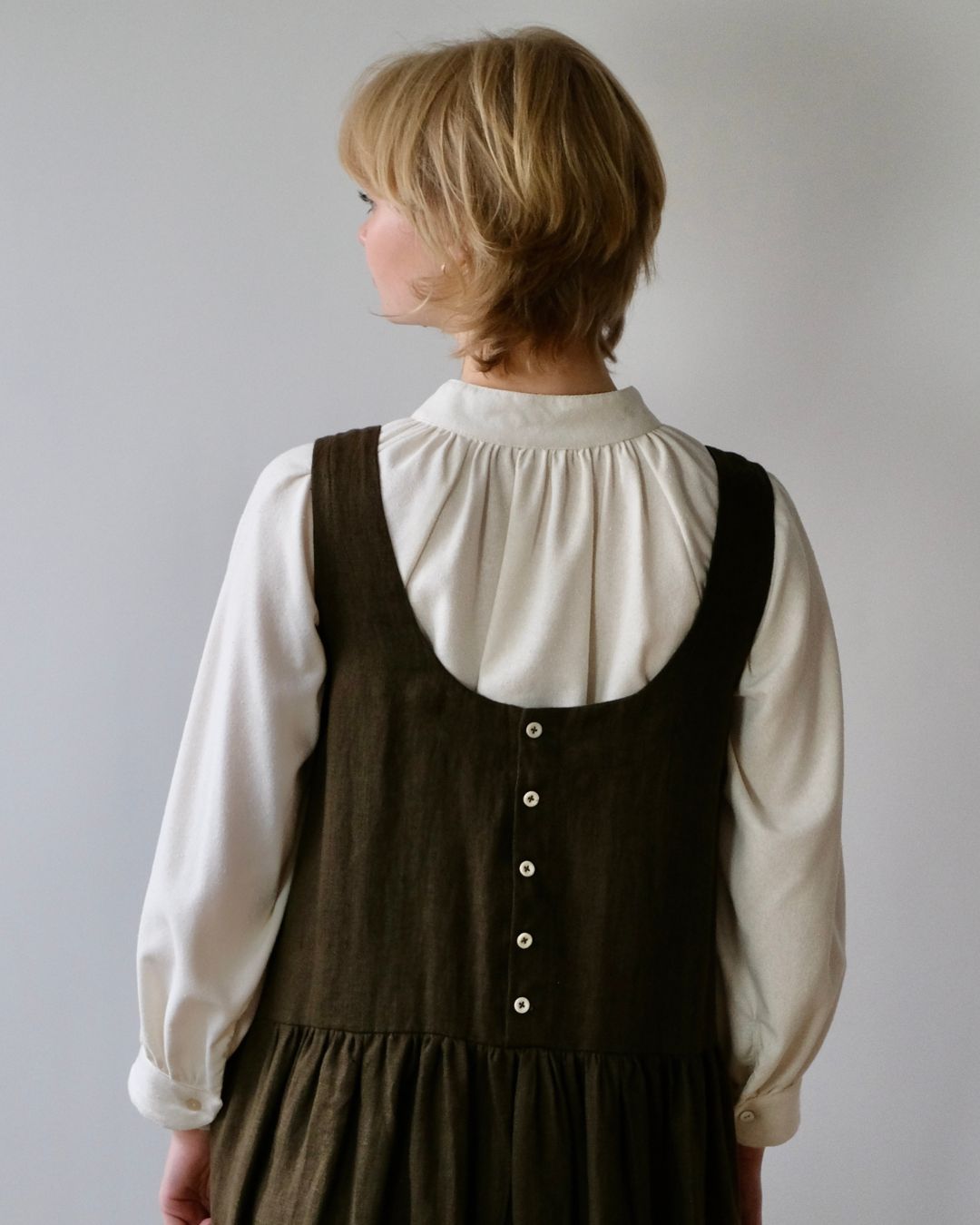 woman standing in conservatory wearing sleeveless pinafore dress and white blouse.