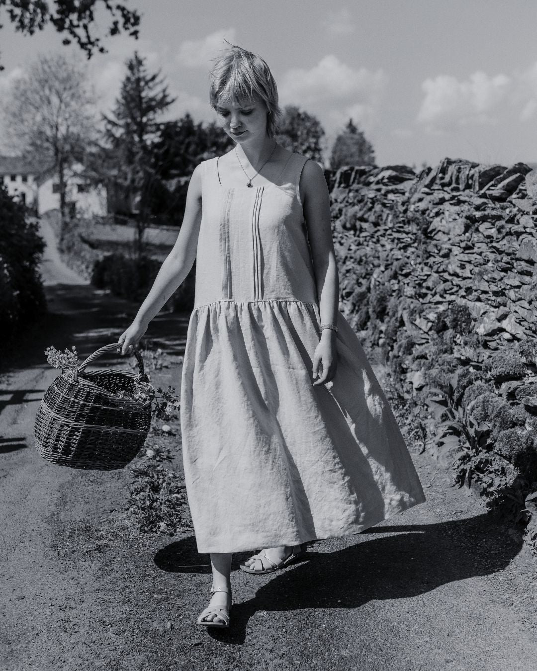 woman outdoors in country wearing sleeveless pinafore dress with pleated front. Farm house in the background.