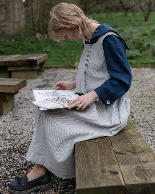 person sat painting in garden wearing stripe pinafore dress and blue shirt
