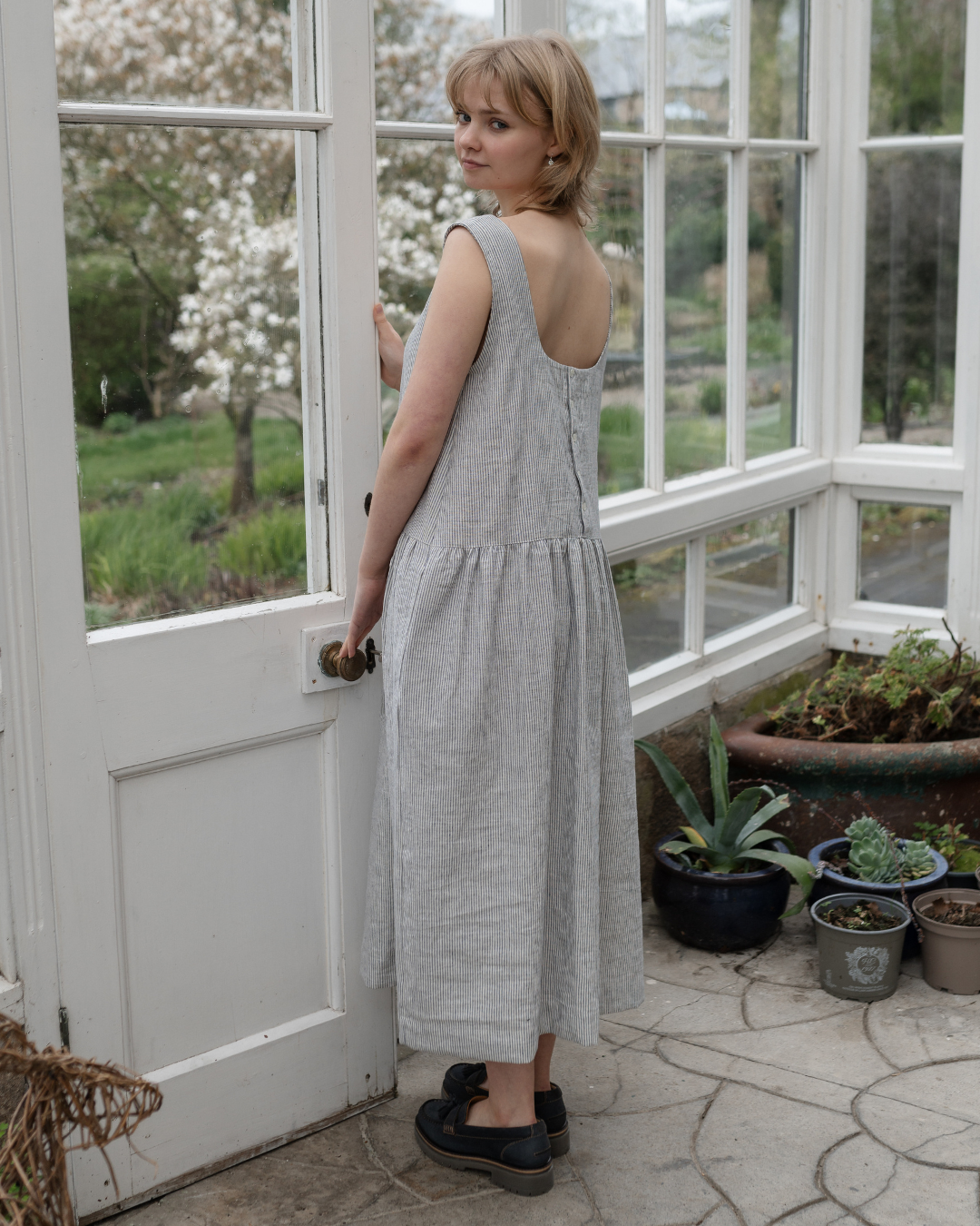 A woman standing in a conservatory wearing stripe linen pinafore dress with pockets
