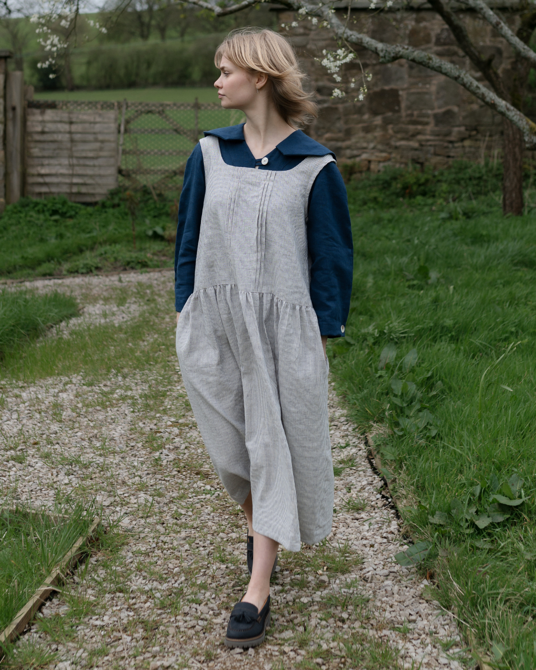 A woman standing in a conservatory wearing a sleeveless, striped, low waisted pinafore dress with a pleated front and side pockets.