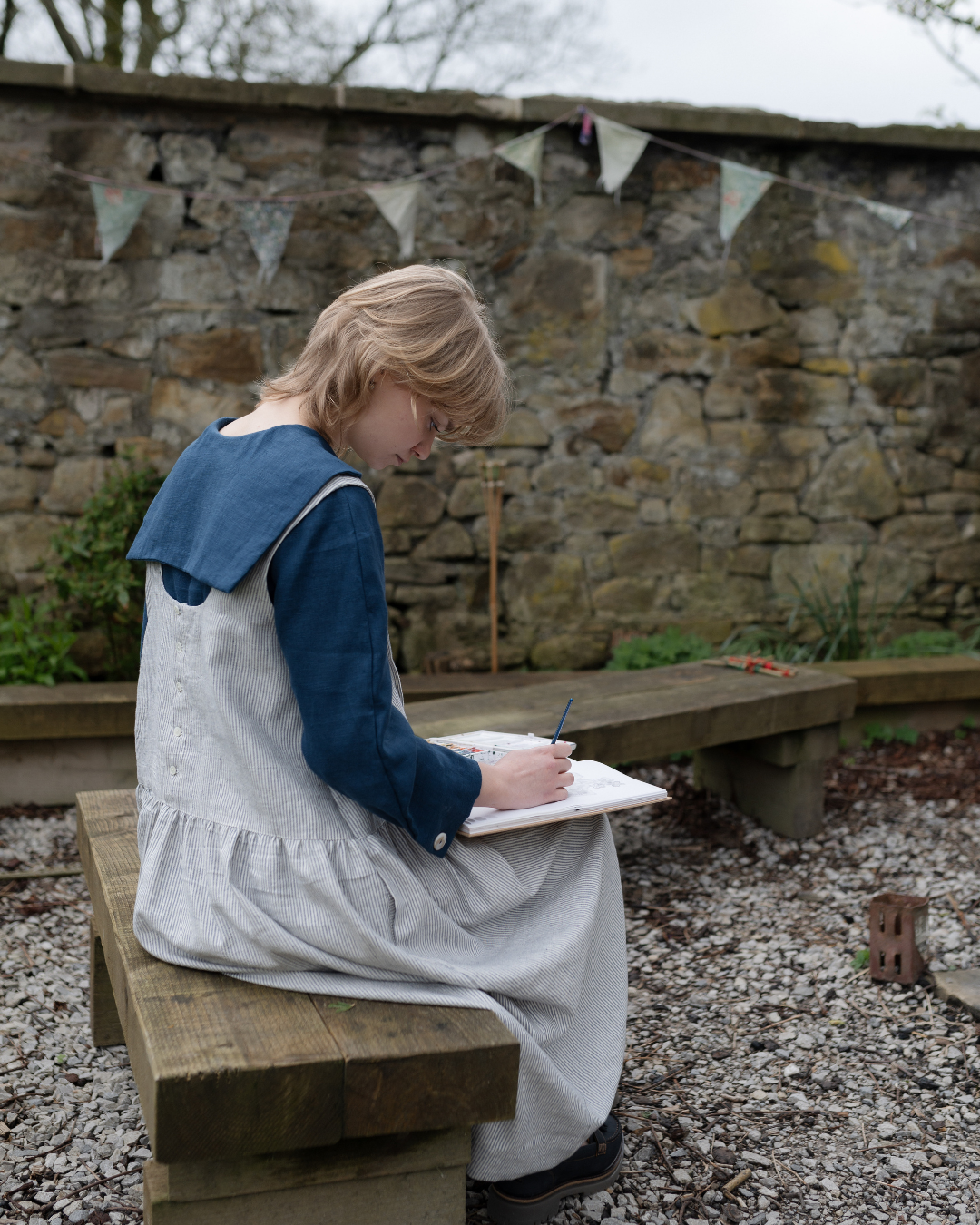 A woman sat in garden drawing wearing stripe pinafore dress with blue shirt