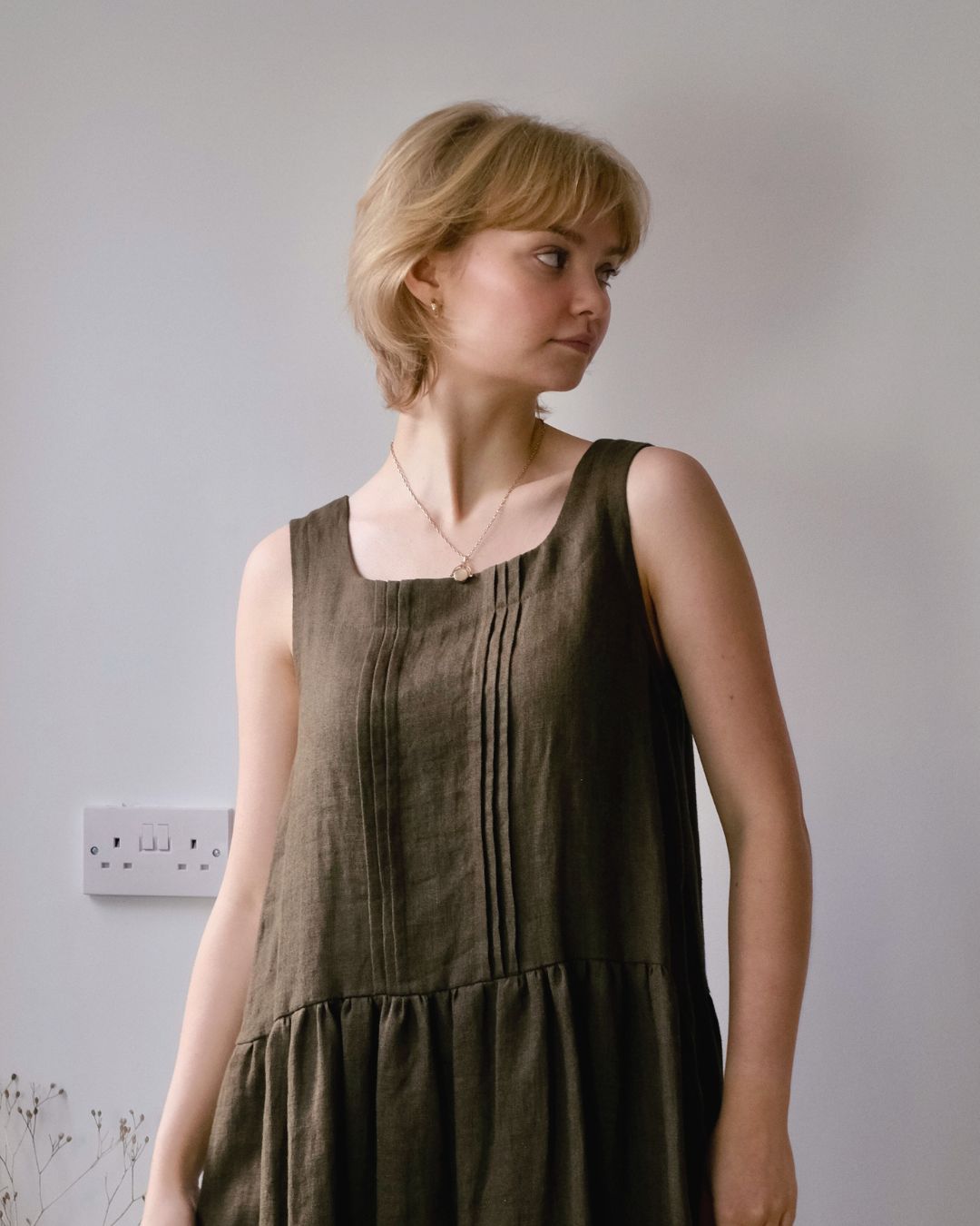 woman standing in conservatory wearing sleeveless pinafore dress with pleated front and pockets.