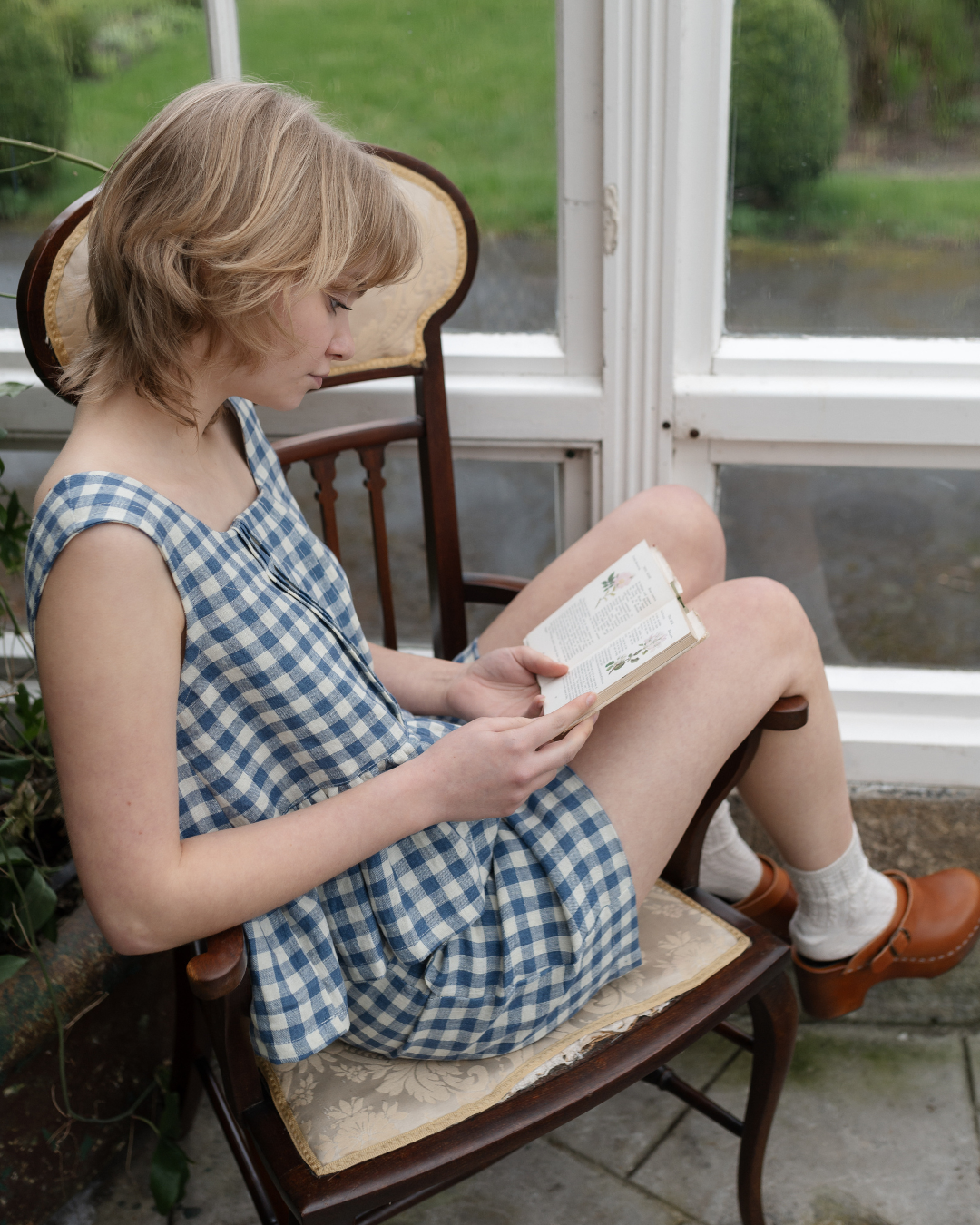 Woman sitting in wooden chair reading a book and wearing blue checkered top and shorts.