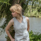 Woman in a light gray sleeveless top and pants standing by a tree and water body.