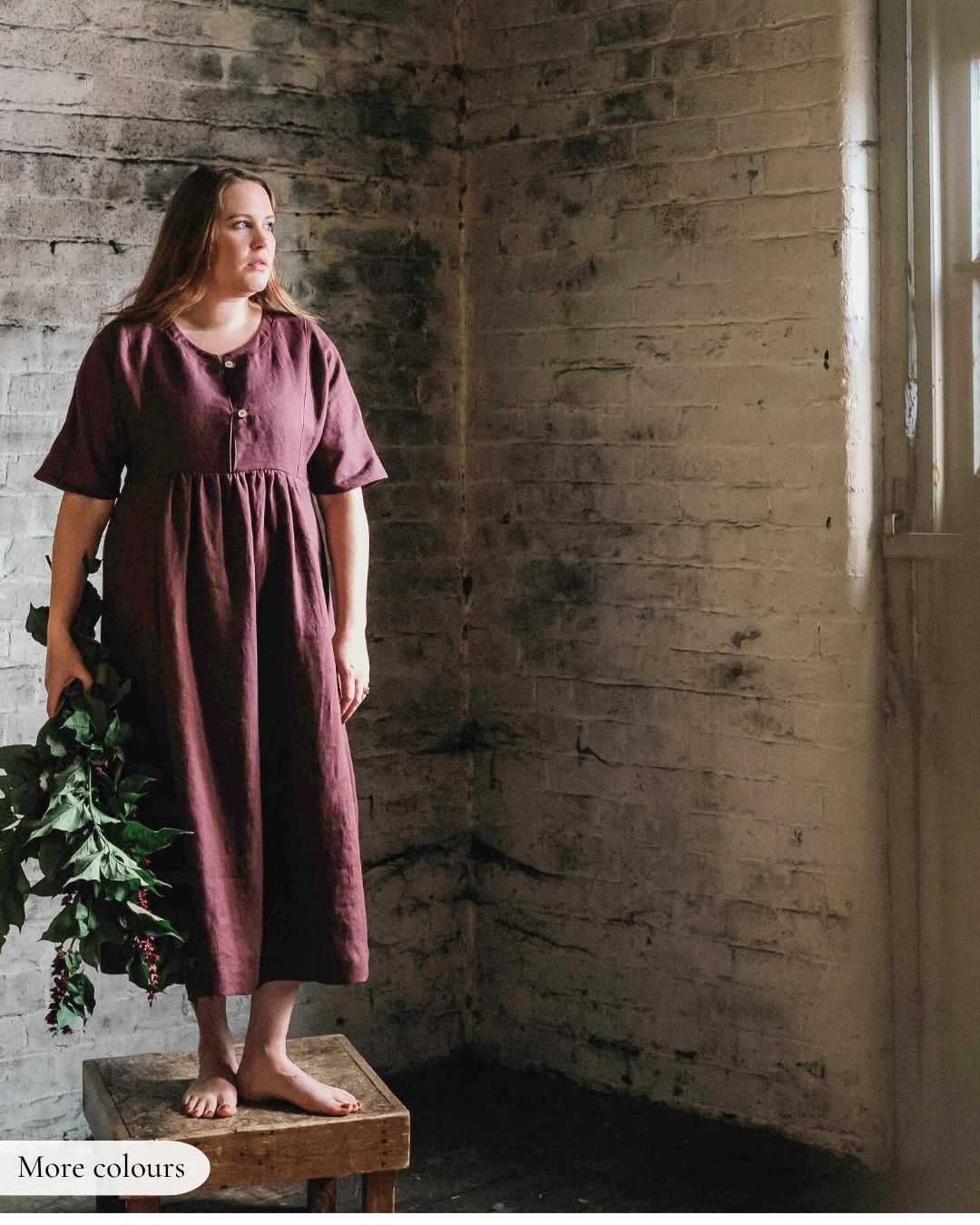 Woman in a maroon dress holding greenery in a rustic room.