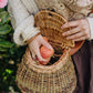 A person holding a traditional wicker basket with a leather strap and wooden fastener, filled with what appears to be foraged items.