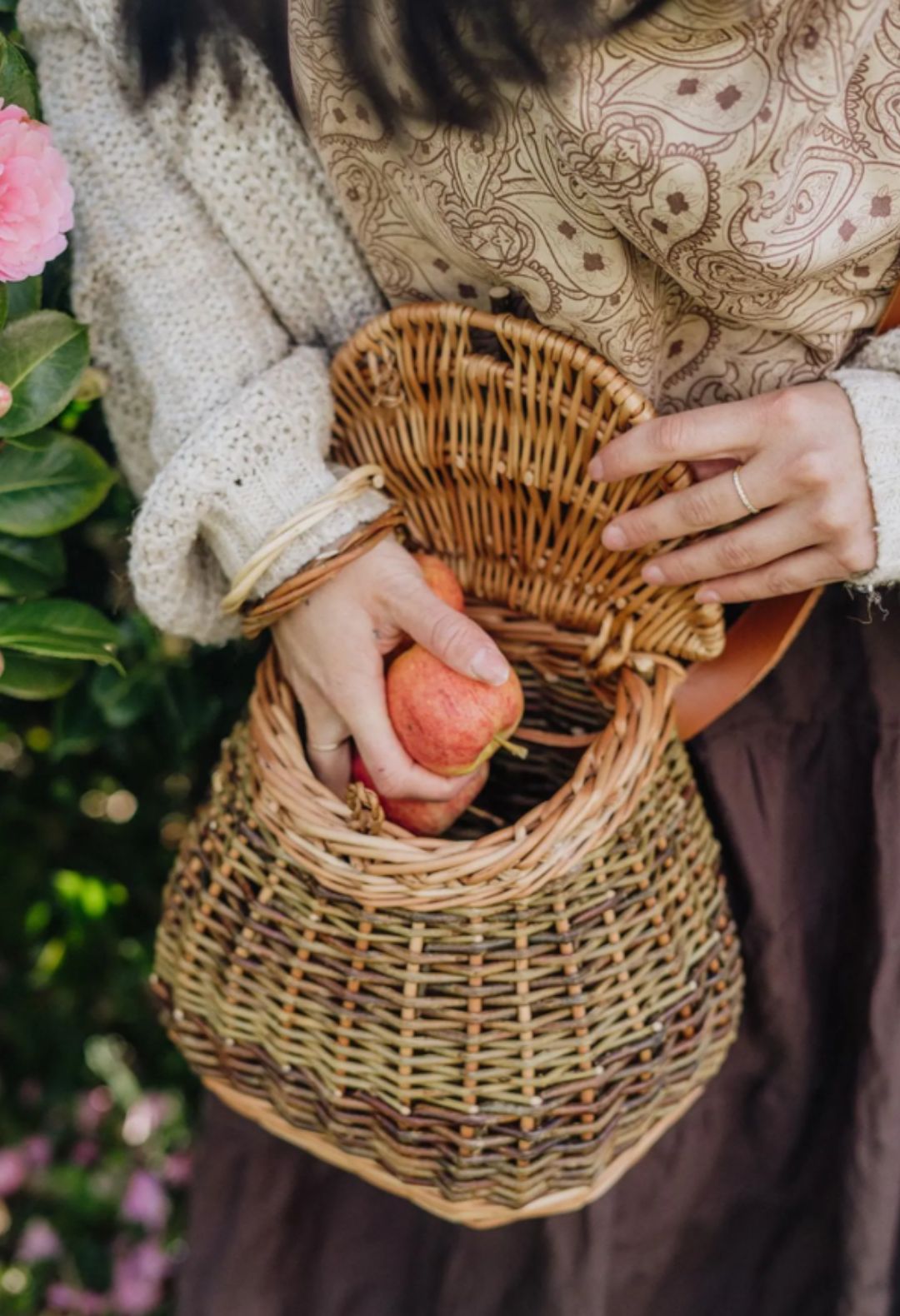 A person holding a traditional wicker basket with a leather strap and wooden fastener, filled with what appears to be foraged items.