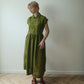 A woman wearing a muted olive green linen midi dress with a buttoned collar and short sleeves, standing on a light wood herringbone floor against a plain off-white wall.