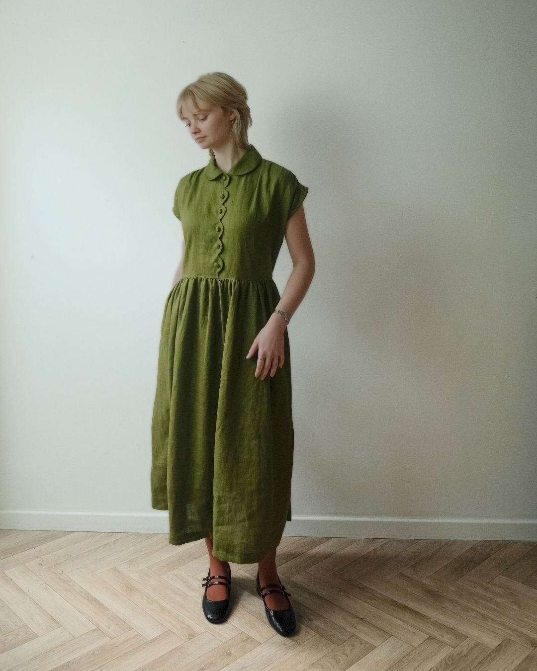 A woman wearing a muted olive green linen midi dress with a buttoned collar and short sleeves, standing on a light wood herringbone floor against a plain off-white wall.
