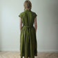 A woman viewed from behind wearing an olive green linen midi dress with a tie waist and short cap sleeves, standing on a light wood herringbone floor against a plain off-white wall.