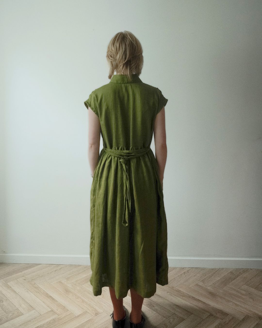 A woman viewed from behind wearing an olive green linen midi dress with a tie waist and short cap sleeves, standing on a light wood herringbone floor against a plain off-white wall.