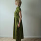 A woman stands in profile wearing a sleeveless olive green midi dress with a gathered waist and black flat shoes, standing on a light wood herringbone floor against a plain off-white wall.