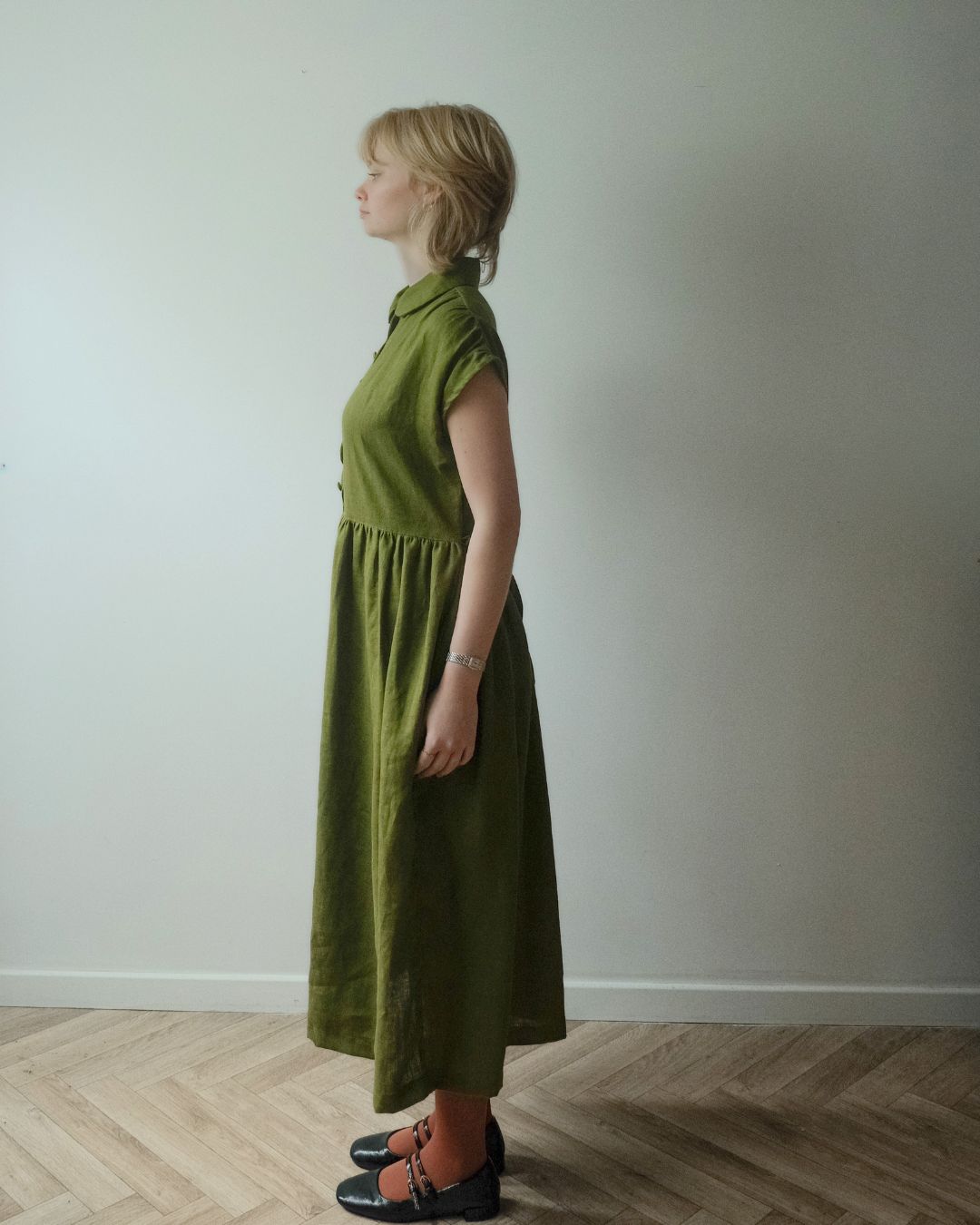 A woman stands in profile wearing a sleeveless olive green midi dress with a gathered waist and black flat shoes, standing on a light wood herringbone floor against a plain off-white wall.