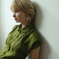 A woman with short blonde hair leans against a white wall, wearing an olive green short-sleeve button-up shirt with a Peter Pan collar.