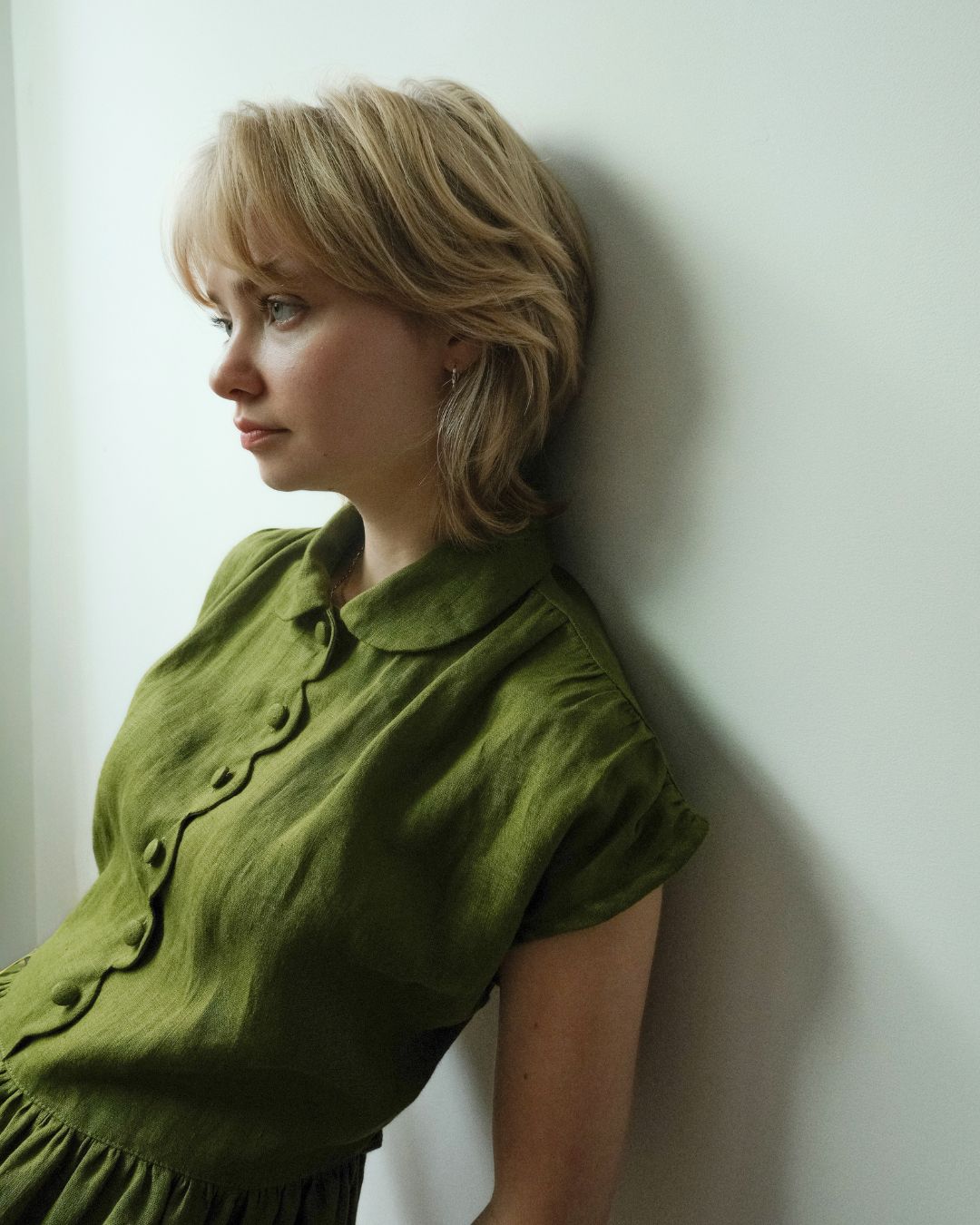 A woman with short blonde hair leans against a white wall, wearing an olive green short-sleeve button-up shirt with a Peter Pan collar.