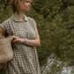 Woman in a checkered dress holding a woven bag, standing by a natural setting with trees and water.